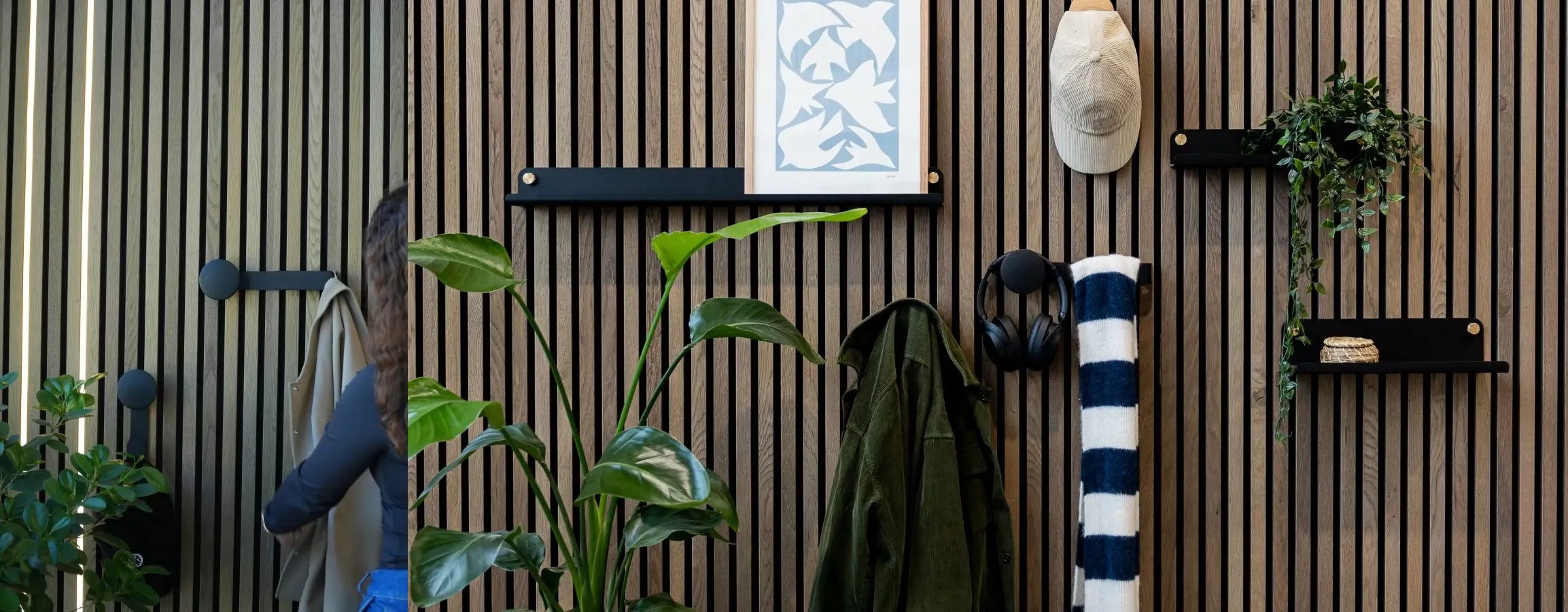 Un uomo in giacca blu passa davanti a una parete acustica nordica in legno Akupanel con piante integrate, mensole, appendiabiti, cuffie e un asciugamano a righe arrotolato.